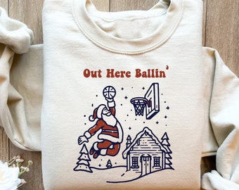 May include: Cream-colored sweatshirt with a graphic of Santa Claus dunking a basketball. The text "Out Here Ballin'" is above the image. The design includes a basketball hoop, a small cabin, and pine trees.