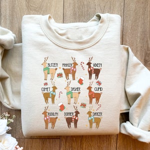 May include: A cream-colored sweatshirt featuring a festive design of nine reindeer, each labeled with a name like "Blitzen" and "Rudolph." The reindeer are depicted with colorful scarves and holiday-themed accents, perfect for Christmas.