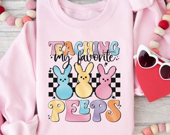 Peut inclure: Sweat-shirt rose avec le texte "TEACHING my favorite PEEPS" en couleurs arc-en-ciel. Trois poussins de dessin animé Peeps sont dans un motif à carreaux derrière le texte.