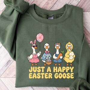 Könnte beinhalten: Ein olivgrünes Sweatshirt mit einer Grafik von vier Cartoon-Gänsen, die für Ostern gekleidet sind. Eine Gans trägt eine Sonnenbrille und ein rosa Kleid und hält einen Ballon. Der Text "JUST A HAPPY EASTER GOOSE" ist gelb gedruckt.