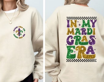 May include: A beige sweatshirt with a purple, green, and gold glitter design on the back that says "In My Mardi Gras Era". The front of the sweatshirt has a small purple, green, and gold glitter design of a fleur-de-lis.
