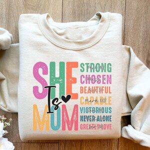 May include: A beige sweatshirt with a colourful graphic print that says "She is Mom" with the words "Strong, Chosen, Beautiful, Capable, Victorious, Never Alone, Greatly Loved" in a rainbow of colours. The text "Mark 10:27" is also included in the graphic.