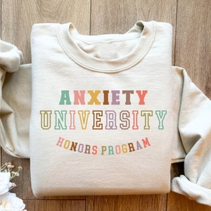 May include: A beige sweatshirt with the text "Anxiety University Honors Program" in colorful letters. The text is in a retro font style.