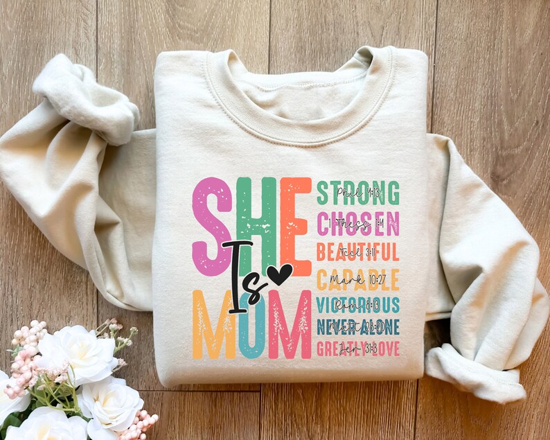 May include: A beige sweatshirt with a colourful graphic print that says "She is Mom" with the words "Strong, Chosen, Beautiful, Capable, Victorious, Never Alone, Greatly Loved" in a rainbow of colours. The text "Mark 10:27" is also included in the graphic.