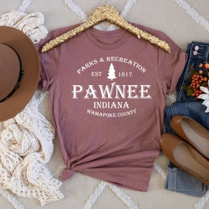 Peut inclure: Un t-shirt mauve avec un motif graphique blanc qui dit "Parks & Recreation Est. 1817 Pawnee Indiana Wapapoke County". Le graphique comprend &eacute;galement un pin stylis&eacute;.