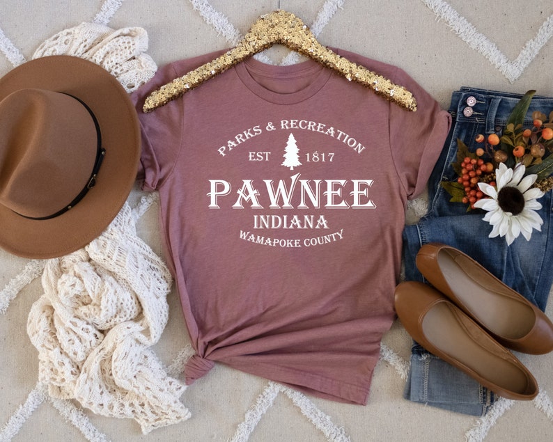 Peut inclure: Un t-shirt mauve avec un motif graphique blanc qui dit "Parks & Recreation Est. 1817 Pawnee Indiana Wapapoke County". Le graphique comprend &eacute;galement un pin stylis&eacute;.