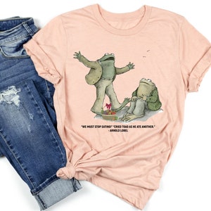Peut inclure: Un t-shirt rose p&acirc;le avec un graphique de deux grenouilles vertes. La grenouille de gauche est debout sur une assiette de nourriture et la grenouille de droite est assise sur un rocher. Le texte sur le t-shirt dit "WE MUST STOP EATING!" "CRIED TOAD AS HE ATE ANOTHER." - ARNOLD LOBEL