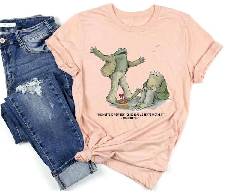 Peut inclure: Un t-shirt rose p&acirc;le avec un graphique de deux grenouilles vertes. La grenouille de gauche est debout sur une assiette de nourriture et la grenouille de droite est assise sur un rocher. Le texte sur le t-shirt dit "WE MUST STOP EATING!" "CRIED TOAD AS HE ATE ANOTHER." - ARNOLD LOBEL