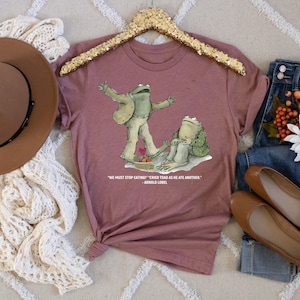 Peut inclure: Un t-shirt mauve avec un graphique de deux grenouilles. Le texte sur le t-shirt dit "On doit arr&ecirc;ter de manger !" cria le crapaud en mangeant un autre. - Arnold Lobel