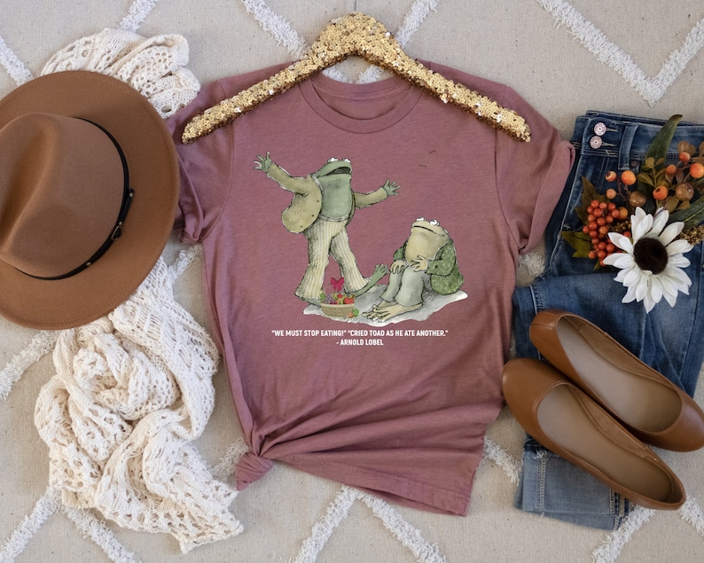 Peut inclure: Un t-shirt mauve avec un graphique de deux grenouilles. Le texte sur le t-shirt dit "On doit arr&ecirc;ter de manger !" cria le crapaud en mangeant un autre. - Arnold Lobel