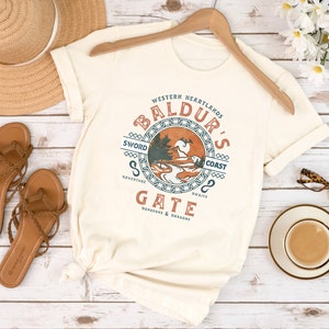 Op de afbeelding: Een wit T-shirt met een vintage-stijl grafisch ontwerp met de tekst "Baldur's Gate" in een cirkel met een kettingrand. De grafiek bevat ook de tekst "Western Heartlands", "Sword Coast", "Adventure", "Swifts" en "Dungeons & Dragons".