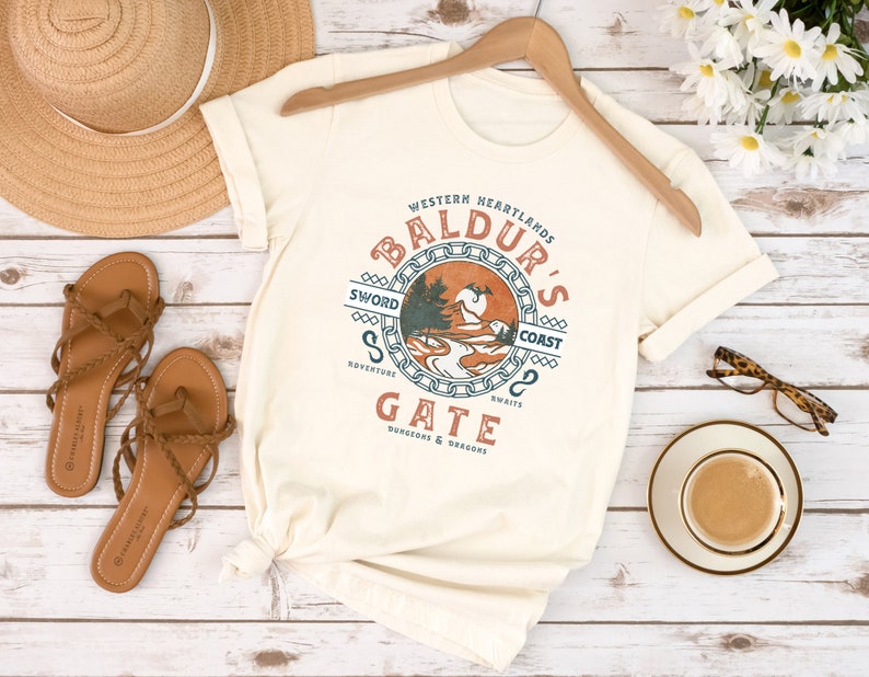 Op de afbeelding: Een wit T-shirt met een vintage-stijl grafisch ontwerp met de tekst "Baldur's Gate" in een cirkel met een kettingrand. De grafiek bevat ook de tekst "Western Heartlands", "Sword Coast", "Adventure", "Swifts" en "Dungeons & Dragons".