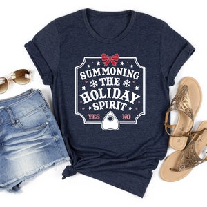 May include: Navy blue t-shirt with the text "SUMMONING THE HOLIDAY SPIRIT" in a white and red design. The shirt includes a planchette graphic and the words "YES" and "NO". The shirt is paired with denim shorts, sunglasses, and sandals.