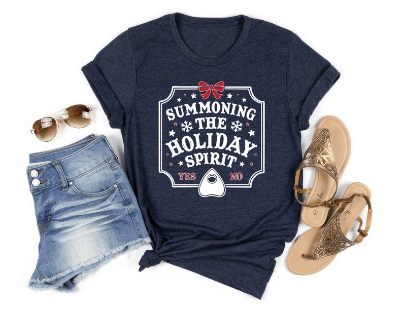 May include: Navy blue t-shirt with the text "SUMMONING THE HOLIDAY SPIRIT" in a white and red design. The shirt includes a planchette graphic and the words "YES" and "NO". The shirt is paired with denim shorts, sunglasses, and sandals.