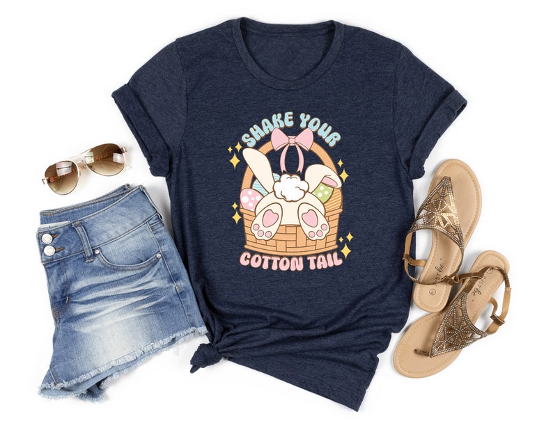 K&ouml;nnte beinhalten: Ein marineblaues T-Shirt mit einem Aufdruck eines Kaninchens in einem Korb mit Eiern. Der Text auf dem Shirt lautet "Shake Your Cotton Tail".