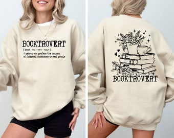 May include: A beige sweatshirt with the text "BOOKTROVERT [book-tro-vert- noun] A person who prefers the company of fictional characters to real people" on the front and a stack of books with a cup of coffee and flowers on the back.