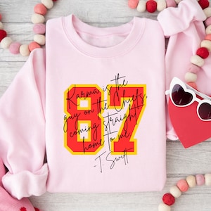 Może przedstawiać: R&oacute;żowa bluza z czerwono-ż&oacute;łtym nadrukiem, na kt&oacute;rym widnieje napis "Karma is the guy on the Chiefs, coming straight home to me. -T. Swift"