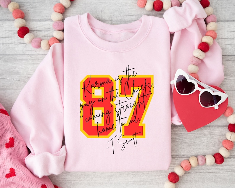 Może przedstawiać: R&oacute;żowa bluza z czerwono-ż&oacute;łtym nadrukiem, na kt&oacute;rym widnieje napis "Karma is the guy on the Chiefs, coming straight home to me. -T. Swift"