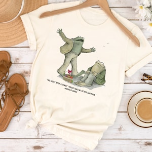 Peut inclure: Un t-shirt cr&egrave;me avec un motif de deux grenouilles. La grenouille de gauche est debout sur une assiette de nourriture et la grenouille de droite est assise. La citation "We must stop eating!" cried Toad as he ate another." - Arnold Lobel est imprim&eacute;e sous le motif.
