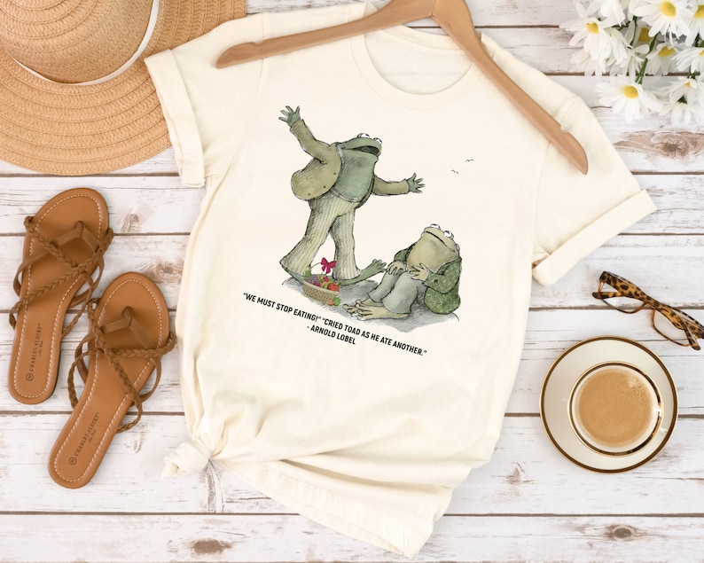 Peut inclure: Un t-shirt cr&egrave;me avec un motif de deux grenouilles. La grenouille de gauche est debout sur une assiette de nourriture et la grenouille de droite est assise. La citation "We must stop eating!" cried Toad as he ate another." - Arnold Lobel est imprim&eacute;e sous le motif.