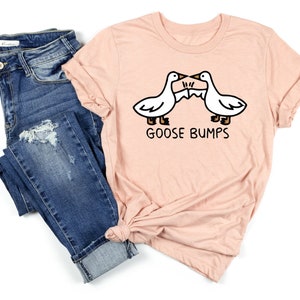May include: A light pink t-shirt with a graphic of two geese facing each other with the text "Goose Bumps" underneath. The shirt is paired with blue jeans.