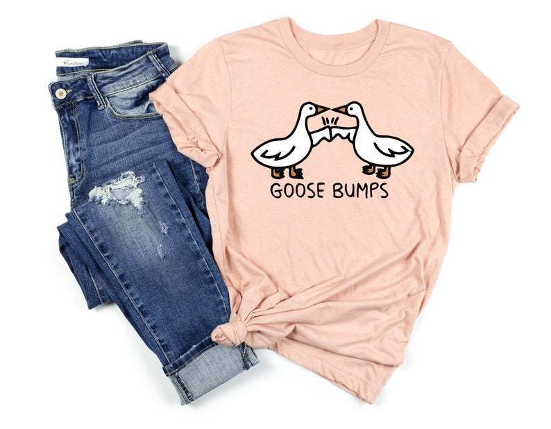 May include: A light pink t-shirt with a graphic of two geese facing each other with the text "Goose Bumps" underneath. The shirt is paired with blue jeans.