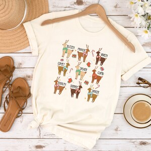 May include: Cream-colored t-shirt featuring cartoon reindeer with names like Blitzen, Comet, and Rudolph. Each reindeer is adorned with festive accessories such as scarves, candy canes, and gift boxes. The shirt is displayed on a wooden hanger.