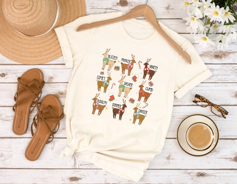 May include: Cream-colored t-shirt featuring cartoon reindeer with names like Blitzen, Comet, and Rudolph. Each reindeer is adorned with festive accessories such as scarves, candy canes, and gift boxes. The shirt is displayed on a wooden hanger.