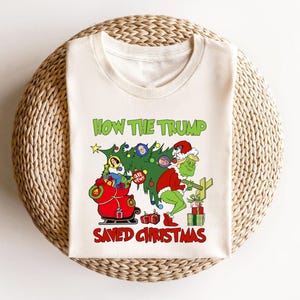 以下が含まれることがあります： 緑色のグリンチのような人物がクリスマスツリーを持っている白いTシャツ。ツリーは、人々の顔が付いたオーナメントで飾られています。Tシャツには「How the Trump Saved Christmas」という文字が印刷されています。
