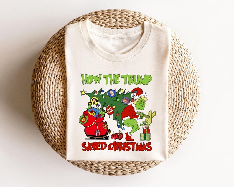 以下が含まれることがあります： 緑色のグリンチのような人物がクリスマスツリーを持っている白いTシャツ。ツリーは、人々の顔が付いたオーナメントで飾られています。Tシャツには「How the Trump Saved Christmas」という文字が印刷されています。