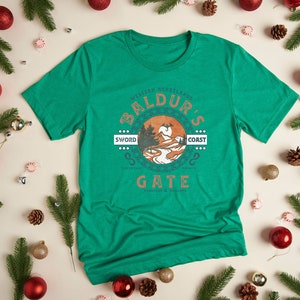 Op de afbeelding: Een groen T-shirt met een vintage-stijl grafisch ontwerp met de tekst "Forgotten Realms Baldur's Gate Sword Coast", een landschapssc&egrave;ne en de tekst "Gate Dungeons & Dragons".