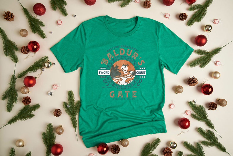 Op de afbeelding: Een groen T-shirt met een vintage-stijl grafisch ontwerp met de tekst "Forgotten Realms Baldur's Gate Sword Coast", een landschapssc&egrave;ne en de tekst "Gate Dungeons & Dragons".