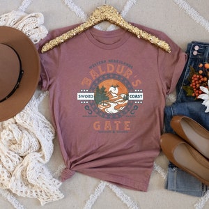 Op de afbeelding: Een mauve t-shirt met een grafische print van een landschap met de tekst "Western Heartlands Baldur's Gate Sword Coast" en een afbeelding van een draak. Het t-shirt hangt aan een hanger met een gouden glitterafwerking.