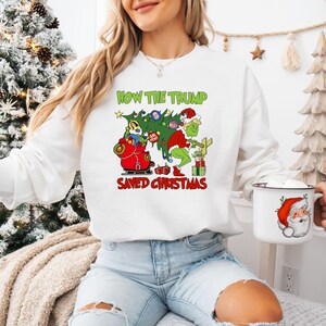 以下が含まれることがあります： クリスマスをテーマにしたグラフィックがプリントされた白いスウェットシャツ。グラフィックには、グリンチとサンタクロースが描かれています。スウェットシャツのテキストは「How the Trump Saved Christmas」です。