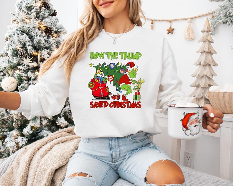 以下が含まれることがあります： クリスマスをテーマにしたグラフィックがプリントされた白いスウェットシャツ。グラフィックには、グリンチとサンタクロースが描かれています。スウェットシャツのテキストは「How the Trump Saved Christmas」です。