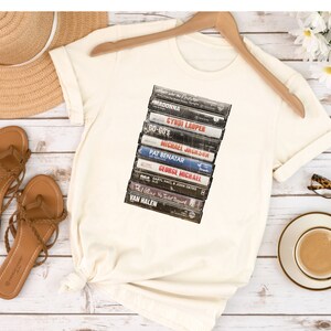 May include: A white t-shirt with a vintage-style graphic of a stack of cassette tapes. The tapes feature the names of popular artists from the 80s, including Madonna, Cyndi Lauper, Michael Jackson, Pat Benatar, George Michael, Van Halen, and more.