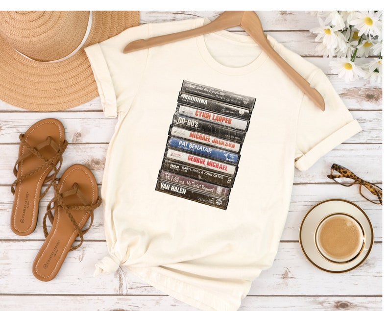May include: A white t-shirt with a vintage-style graphic of a stack of cassette tapes. The tapes feature the names of popular artists from the 80s, including Madonna, Cyndi Lauper, Michael Jackson, Pat Benatar, George Michael, Van Halen, and more.