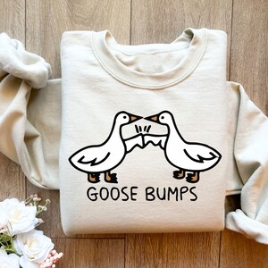May include: A light beige sweatshirt with a graphic of two cartoon geese facing each other with the text "GOOSE BUMPS" printed below.