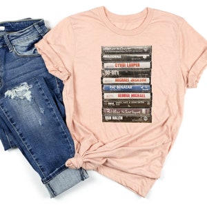 May include: A pink t-shirt with a graphic of a stack of vintage cassette tapes featuring artists such as Madonna, Cyndi Lauper, Go-Go's, Michael Jackson, Pat Benatar, George Michael, Phil Collins, and Van Halen.