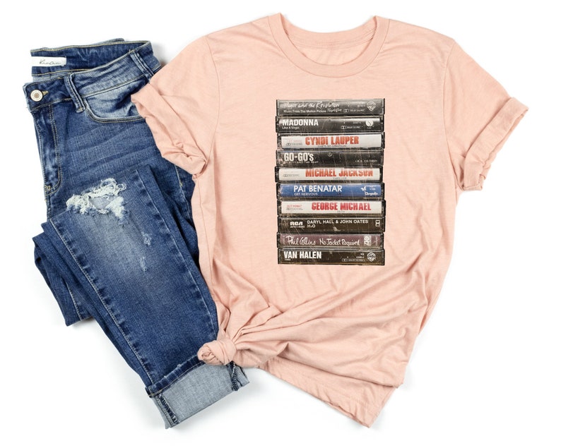 May include: A pink t-shirt with a graphic of a stack of vintage cassette tapes featuring artists such as Madonna, Cyndi Lauper, Go-Go's, Michael Jackson, Pat Benatar, George Michael, Phil Collins, and Van Halen.