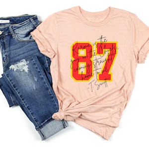Może przedstawiać: R&oacute;żowy t-shirt z czerwono-ż&oacute;łtym nadrukiem, na kt&oacute;rym widnieje napis "Karma is the guy on the Chiefs, coming straight home to me - T. Swift".