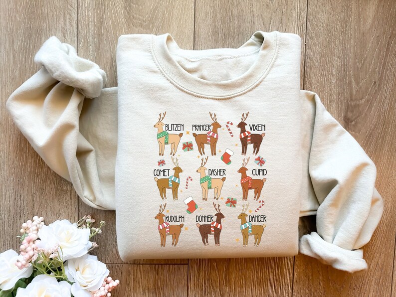 May include: A cream-colored sweatshirt featuring a festive design of nine reindeer, each labeled with a name like "Blitzen" and "Rudolph." The reindeer are depicted with colorful scarves and holiday-themed accents, perfect for Christmas.