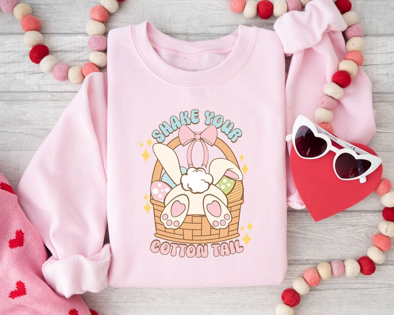 K&ouml;nnte beinhalten: Rosafarbener Pullover mit einem Aufdruck eines Hasen in einem Korb mit Eiern und dem Text "Shake Your Cotton Tail".