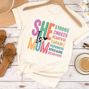 May include: A white t-shirt with the text "She is Mom" in rainbow colours. The text is layered with words like "strong", "chosen", "beautiful", "capable", "victorious", "never alone", and "greatly loved".