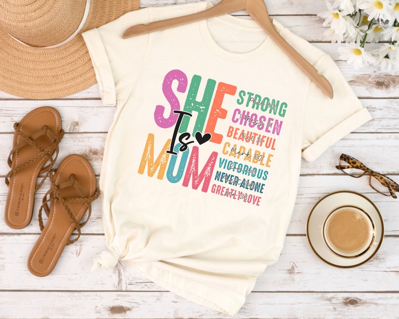 May include: A white t-shirt with the text "She is Mom" in rainbow colours. The text is layered with words like "strong", "chosen", "beautiful", "capable", "victorious", "never alone", and "greatly loved".