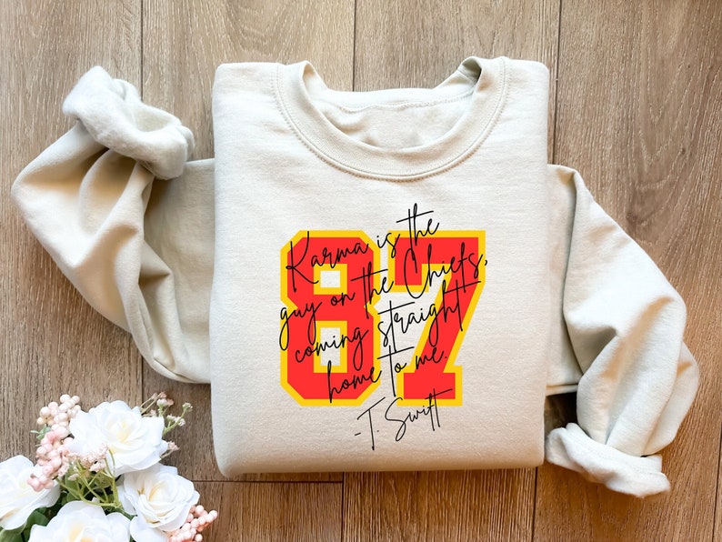 Może przedstawiać: Beżowa bluza z czerwono-ż&oacute;łtym nadrukiem graficznym, na kt&oacute;rym widnieje napis "Karma is the guy on the Chiefs, coming straight home to me - T. Swift".