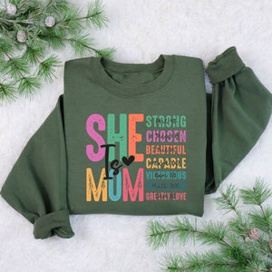 May include: A green sweatshirt with the text "She Is Mom" in colourful letters. The text is surrounded by other words in smaller letters, including "strong", "chosen", "beautiful", "capable", "victorious", "Matt 28:20", and "greatly love".