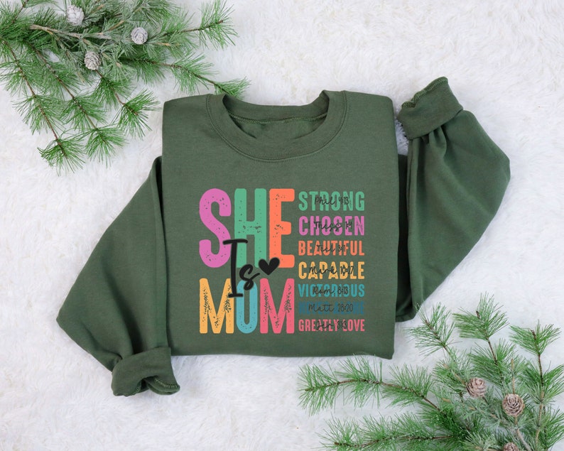 May include: A green sweatshirt with the text "She Is Mom" in colourful letters. The text is surrounded by other words in smaller letters, including "strong", "chosen", "beautiful", "capable", "victorious", "Matt 28:20", and "greatly love".