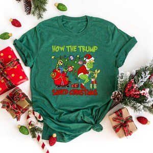 以下が含まれることがあります： 緑色のTシャツに、グリンチの漫画の画像と「How the Trump Saved Christmas」というテキストが書かれています。