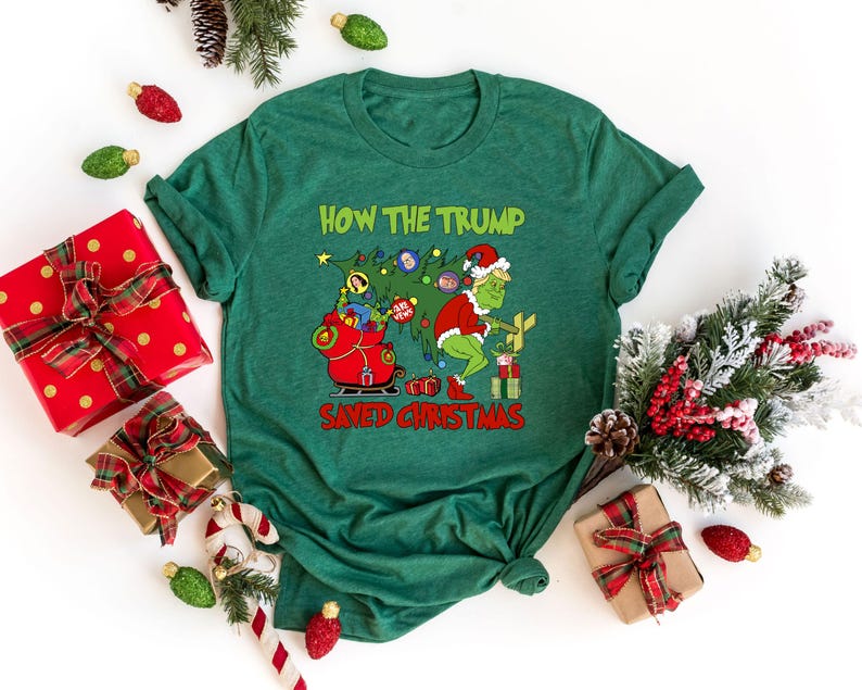 以下が含まれることがあります： 緑色のTシャツに、グリンチの漫画の画像と「How the Trump Saved Christmas」というテキストが書かれています。